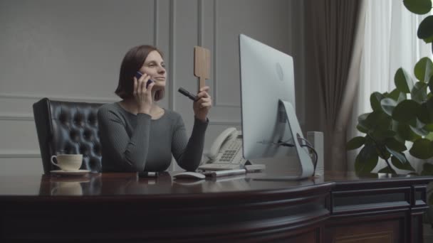 Młoda bizneswoman aplikująca uzupełnić patrząc w lustro w miejscu pracy. Brunetka w biurze biznesowym rozmawia przez smartfona. Biurko z monitorem. — Wideo stockowe