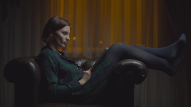 30代の女性がタブレットコンピューターを使って夜な夜な座り心地の良いアームチェアに座り. — ストック動画