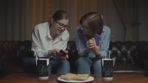 Dos amigas en gafas con smartphones sonriendo y hablando sentadas en un sofá a altas horas de la noche. Caja de comida asiática con palillos en la mesa . — Vídeo de stock