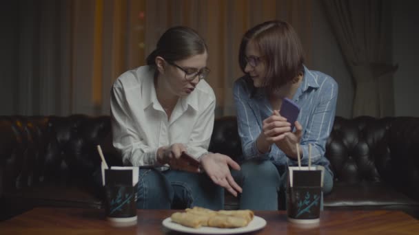 Duas amigas de óculos com smartphones sorrindo e conversando sentadas no sofá tarde da noite. Asiático caixa de comida com pauzinhos na mesa . — Vídeo de Stock