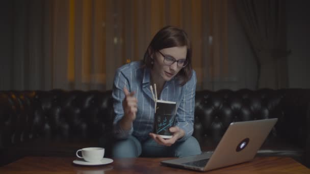 Mujer de 30 años en gafas comiendo comida caja asiática con palillos mirando monitor portátil. Cena sola en casa tarde con gadgets . — Vídeos de Stock