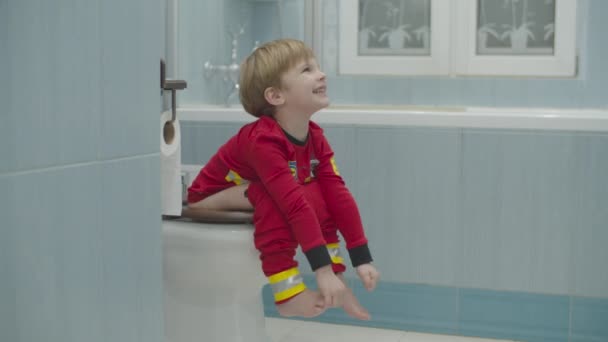 Niño rubio preescolar en pijama de bombero empujando sentado en el inodoro en el baño azul. Niño en el inodoro . — Vídeos de Stock