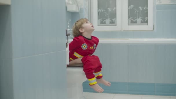 穿着消防员睡衣的金发学龄前男孩坐在蓝色浴室的马桶上推挤着。上厕所的小孩. — 图库视频影像