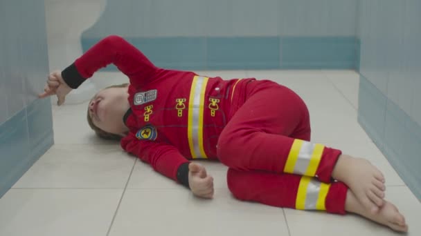 Blonder Vorschuljunge im Feuerwehrschlafanzug, der Spaß auf dem Fußboden des Badezimmers hat. Kind albert auf weißem Boden herum. — Stockvideo