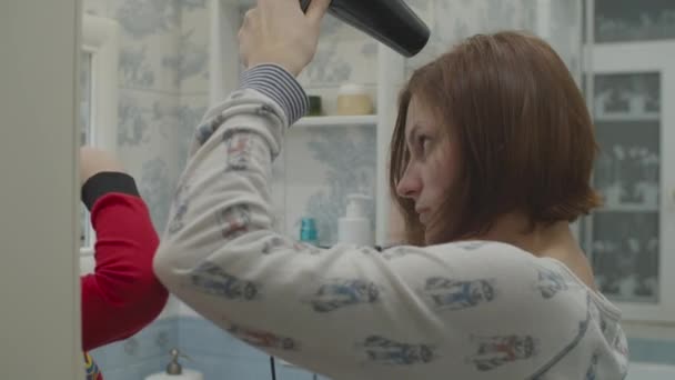 Młoda matka suszy włosy suszarką do włosów. Dzieciak w czerwonej piżamie siedzi obok mamy. Zabawa w rodzinną kąpiel. Mama i syn śmieją się w łazience. — Wideo stockowe
