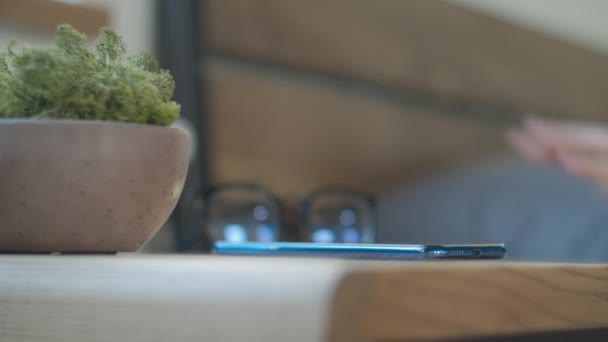 Womans hand touching mobile phones screen to snooze alarm clock in bedroom, taking off glasses from bedside table with moss plant. — Stok video