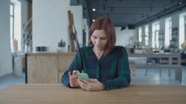 Giovane lavoratrice degli anni '30 che naviga online al cellulare seduta al tavolo di legno in una grande stanza con finestre. Donna che si tocca i capelli . — Video Stock