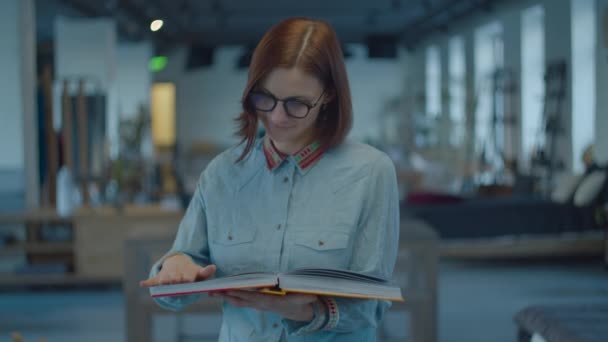 Young 30s female student in ethno shirt and glasses standing with art album in hands. Woman turning pages in album and reading. — Stockvideo