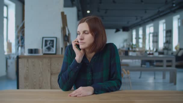 Young 30s female worker talking by mobile phone, sitting at the wooden table in large room with windows. Brunette woman talking holding cell in hands. — Stock Video