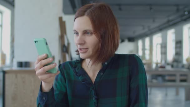 Primer plano de la joven trabajadora de 30 años que habla con la pantalla del teléfono móvil, sentada en la mesa de madera en una habitación grande con ventanas. Morena mujer hablando celebración de la celda en las manos . — Vídeos de Stock