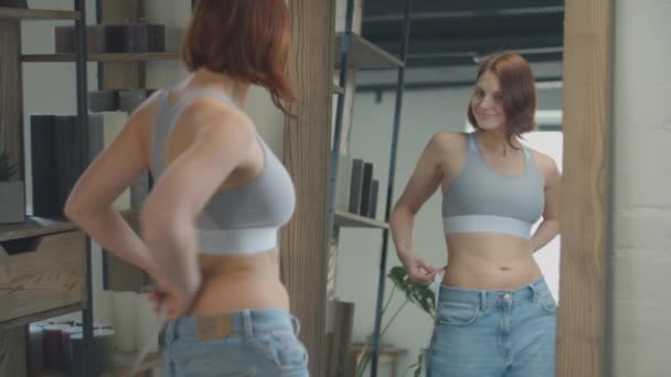 Jonge dertiger jaren vrouw bewondert het resultaat van gewichtsverlies tijdens het dragen van grote maat oude jeans. gelukkig vrouw glimlachen in spiegel met centimeter tape — Stockvideo