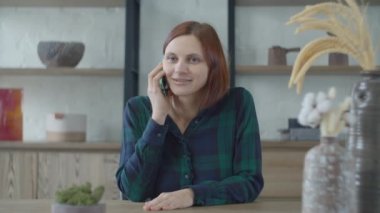 Young smiling 30s female in green dress talking by mobile phone sitting at the table with ikebana vase. Woman laughing during phone talk. 