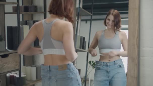 Young 30s woman admiring the result of weight loss while wearing big size old jeans. Happy female smiling in mirror with centimeter tape — 图库视频影像