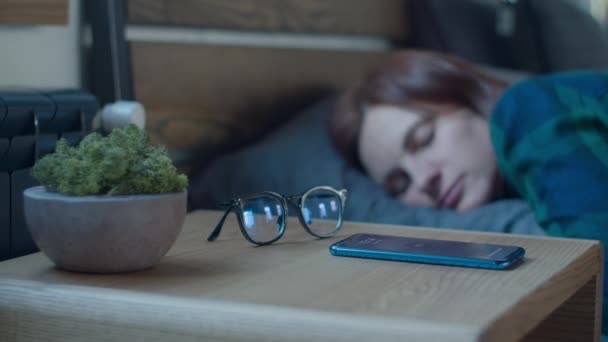 Sleepy female waking up with alarm clock on mobile phone on bedside table with moss plant and glasses. Womans hand touching cell screen to snooze alarm clock in bedroom. — ストック動画