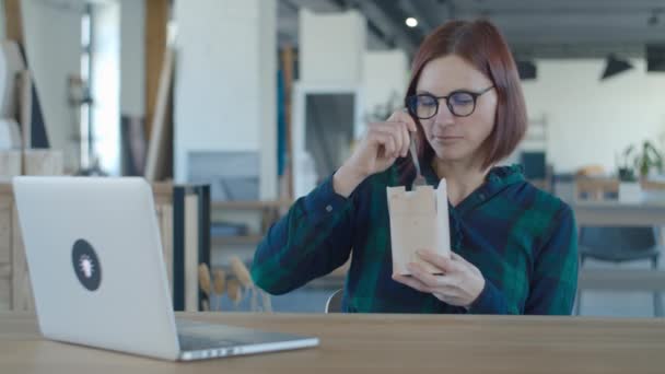 Głodna pracownica jedząca lunch z papierowego pudełka i oglądająca zabawne treści na laptopie. Kobieta je makaron na biurku z laptopem. — Wideo stockowe