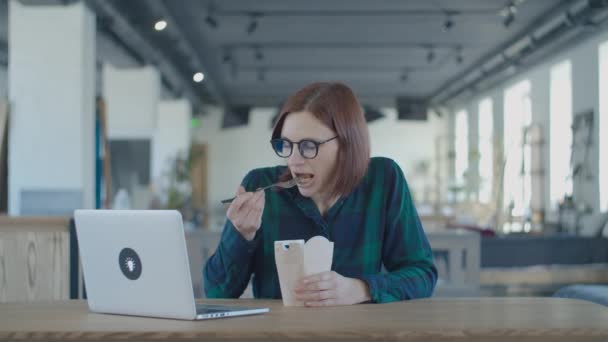 Голодна жінка-працівник їсть обід з паперової коробки і дивиться смішний контент на ноутбуці. Жінка їсть локшину за столом з ноутбуком . — стокове відео