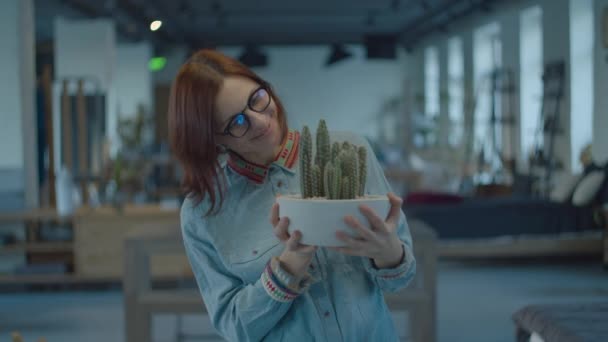 Jonge lachende dertiger jaren vrouw in etno shirt en bril staan met potted cactus plant in handen. Vrouw die rond kijkt naar cactus in pot en glimlacht. — Stockvideo