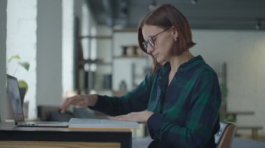 Young female adult student in glasses sitting at the desk with album and laptop and studying. Woman looking to book and typing on laptop. 