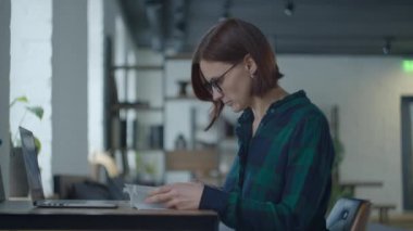 Young female adult student in glasses sitting at the desk with book and laptop and studying. Woman reading book and typing on laptop. 