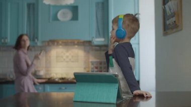 Blonde preschooler boy in color headphones using tablet computer at the table on the kitchen. Mother comes for help 