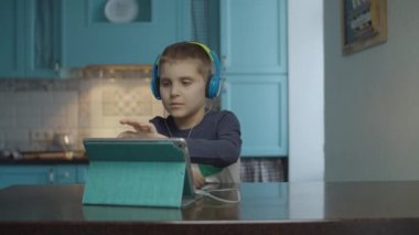 Autistic boy using tablet computer with headphones on the kitchen. Kid with autism looking around at home. Autism awareness