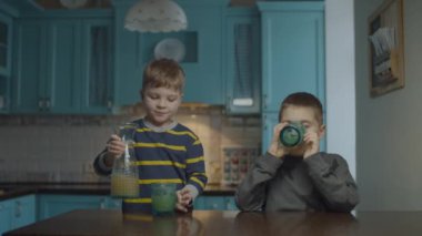 Two boys drinking apple juice from glasses on kitchen. Younger brother pouring juice from carafe into glasses. Siblings at home. 