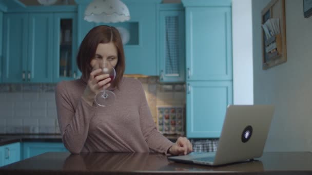 Jeune femme souriante des années 30 buvant un verre de vin rouge et tapant sur l'ordinateur de la cuisine. Femme avec vin et gadget à la maison . — Video
