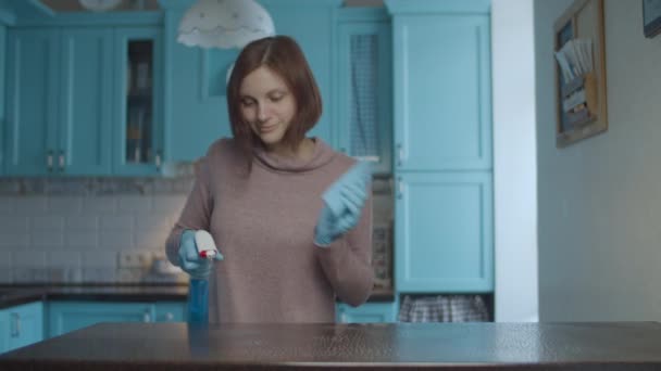 Молодая женщина 30-х годов распыляет моющее средство и вытирает тряпкой поверхность стола. Танцующая и улыбающаяся женщина, убирающая дом . — стоковое видео