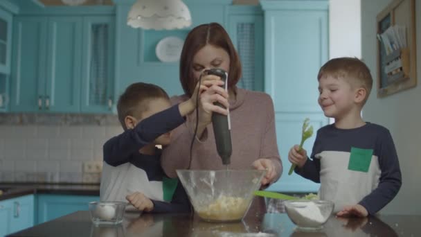 Діти допомагають матері замішувати тісто блендером. Щаслива сім'я готує печиво і розважається на синій домашній кухні . — стокове відео