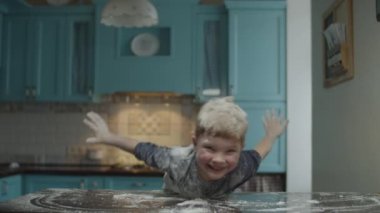 Blonde preschooler having fun with flour on blue kitchen. Boy playing on table with flour. Kid pretending to be airplane. 