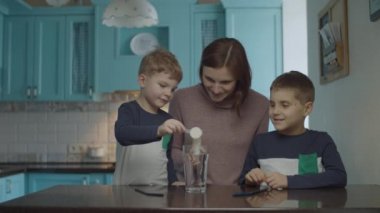 Young mother doing homemade soda drink with two kids on the kitchen. Kids drinking beverage with bubbles through drinking straw. 