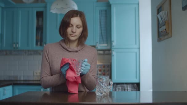 Joven mujer de 30 años sonriente con guantes limpiando tazas de vidrio con servilleta rosa y bailando en la cocina azul. Mujer feliz con la limpieza . — Vídeos de Stock