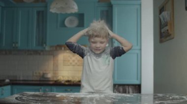 Blonde preschooler having fun with flour on blue kitchen. Boy playing on table with flour. Kid sprinkling his head with flour and smiling. 