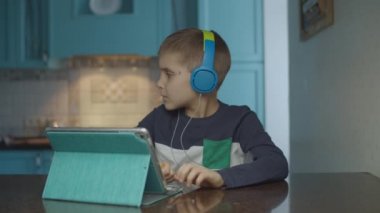 Autistic boy using tablet computer with headphones on the kitchen. Kid with autism looking around at home. Autism awareness