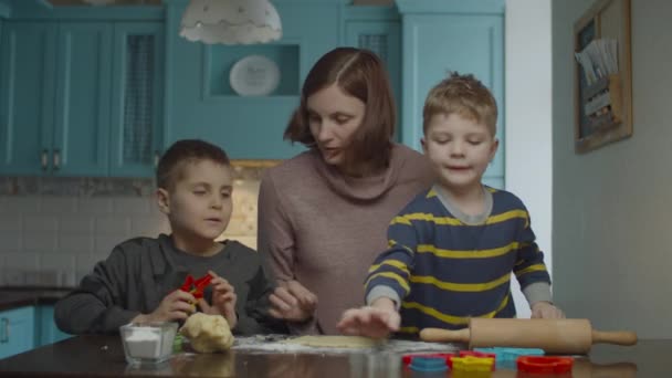 Madre joven y dos hijos cocinando diferentes formas de galletas de masa. Niños ayudando a mamá a cocinar dulces en casa cocina azul . — Vídeo de stock