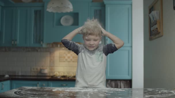 Blonde preschooler having fun with flour on blue kitchen. Boy playing on table with flour. Kid sprinkling his head with flour and smiling. — Stok video