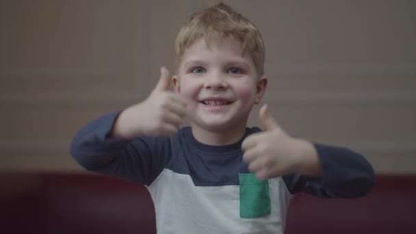Portret blondyna z przedszkola uśmiechającego się i pokazującego kciuki do góry, patrzącego w kamerę. Młody pozytywny dzieciak ściskający dłonie z kciukami w górze. — Wideo stockowe