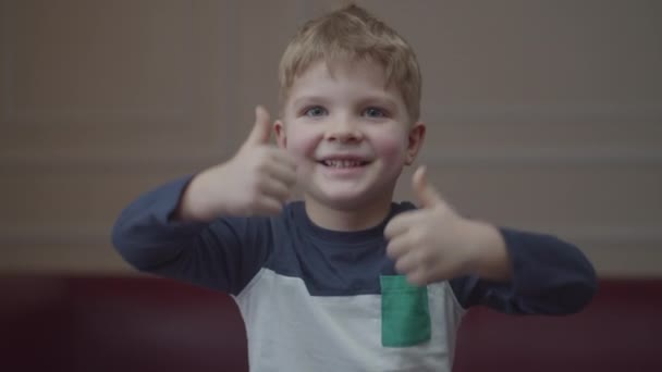 Retrato de niño rubio preescolar sonriendo y mostrando los pulgares hacia arriba, mirando a la cámara. Joven niño positivo estrechando las manos con los pulgares hacia arriba . — Vídeo de stock