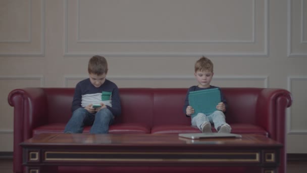 Due fratelli con gli stessi vestiti che usano gadget sul divano di casa. Fratelli che guardano diversi dispositivi seduti sul divano rosso . — Video Stock