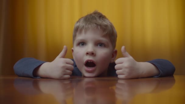 Portret van een blonde kleuter die glimlacht en naar de camera kijkt. Kinderen gaan thuis op de tafel liggen. Glimlachend kind toont duimen omhoog. — Stockvideo