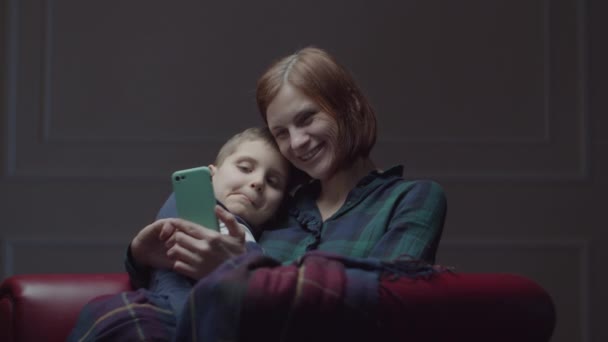 Jeune mère des années 30 avec un fils écolier utilisant un téléphone mobile, assis sur un fauteuil confortable. Famille s'amuser avec gadget à la maison . — Video