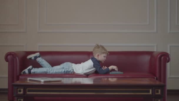 Blond förskolekille med surfplatta, liggandes på soffan hemma. Grabben med gadget ligger och svingar benen på röd soffa. — Stockvideo
