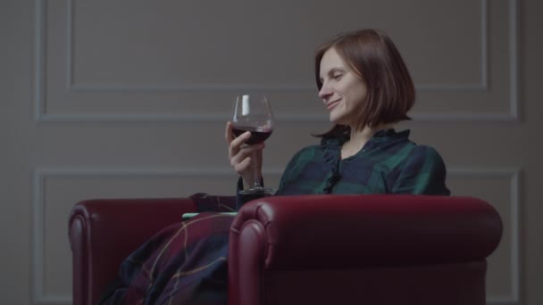 Giovane 30s femminile bere vino rosso seduto in accogliente poltrona rossa a casa. Donna sola con cellulare e bicchiere di vino . — Video Stock