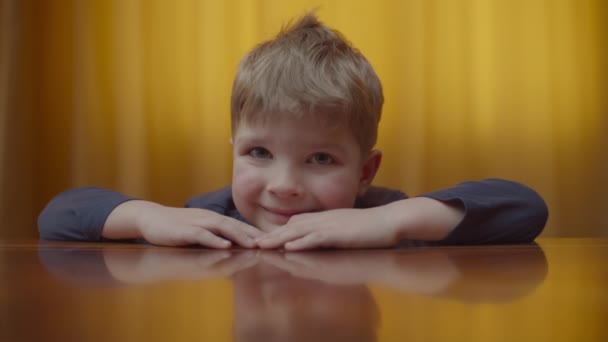 Egy szőke óvodás fiú portréja, aki mosolyog és a kamerába néz. A gyerekek otthon ülnek az asztalon. Mosolygó kölyök felmutatja a hüvelykujját.. — Stock videók