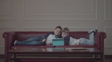 Aynı kıyafetleri giymiş iki kardeş evde kanepede yatıyordu. Kardeşler kanepede bir tablet bilgisayarı izliyor.. 