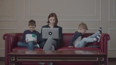 Dizüstü bilgisayarı olan 30 'lu yaşlarda bir anne ve aynı kıyafetleri giyen iki çocuk. Alet kullanıyorlar, kanepede oturuyorlar. Aile, evdeki farklı cihazlardan çevrimiçi içeriği izliyor. 