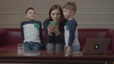 30 'lu yaşlarda bir anne koltukta oturuyor ve cep telefonu kullanıyor. İki oğlu da anneleriyle birlikte akıllı telefon izliyor. Aile evde çevrimiçi içeriği izliyor. 