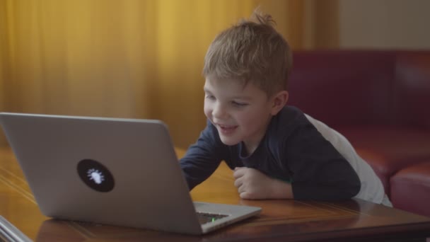 Blonder Vorschuljunge tippt auf Laptop und schaut lächelnd auf Computerbildschirm. Kinder lernen online zu Hause am Tisch. — Stockvideo
