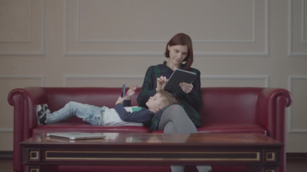 Joven madre de 30 años con tableta y niño acostado en su pierna usando gadget en el sofá. Familia viendo contenido en línea en diferentes dispositivos en casa . — Vídeos de Stock