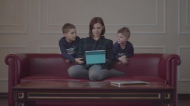 30 'lu yaşlarda genç bir anne kanepede oturuyor ve tablet bilgisayar kullanıyor. Aynı kıyafetleri giymiş iki çocuk annemle birlikte cihaz izliyor. Aile evde çevrimiçi içeriği izliyor. 